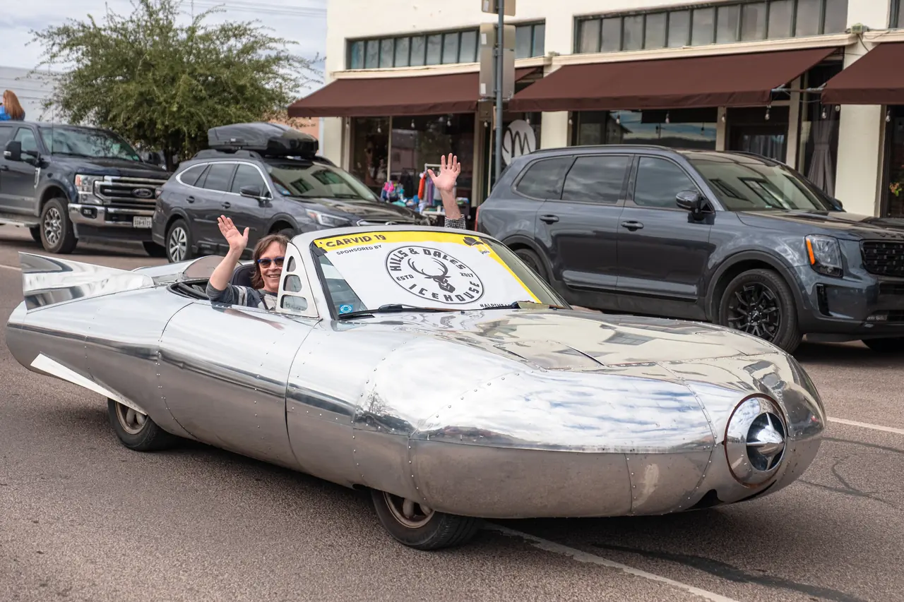 A streamlined polished aluminum convertible art car with tail fins drives down a street while the driver waves.