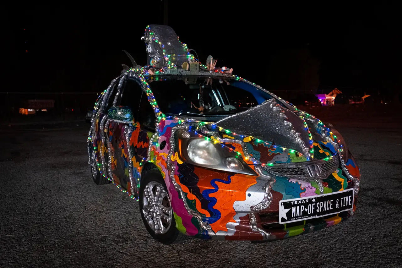 A hatchback painted in swirling colors and wrapped in foil and string lights sits in a dark lot with a sculpted silver fin on its roof.