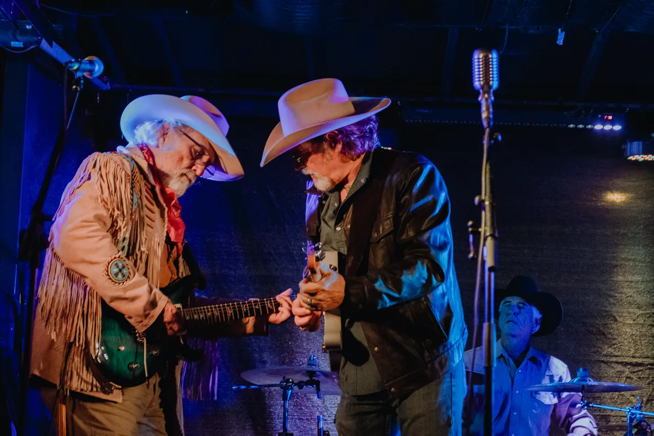 Two guitarists in cowboy hats play together on a stage lit in blue.