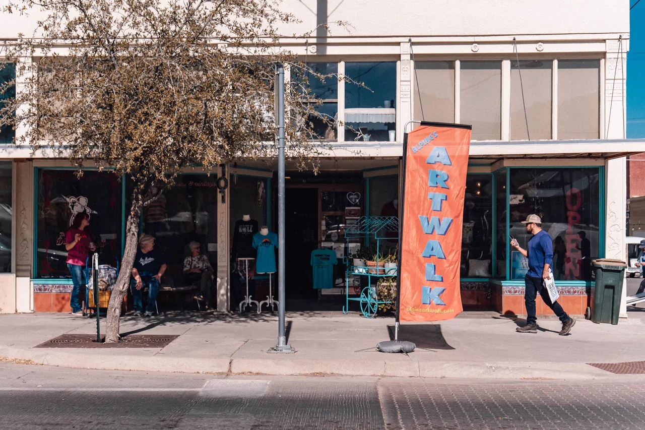 A downtown Alpine storefront with an orange Artwalk banner on the sidewalk and people walking by.