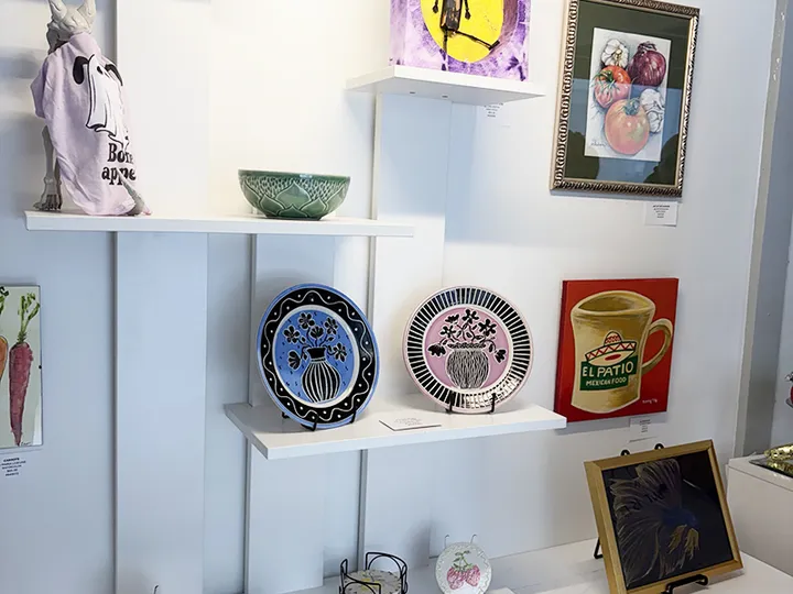 Painted ceramic plates, a bowl and small paintings on white shelves at Big Bend Art Gallery.